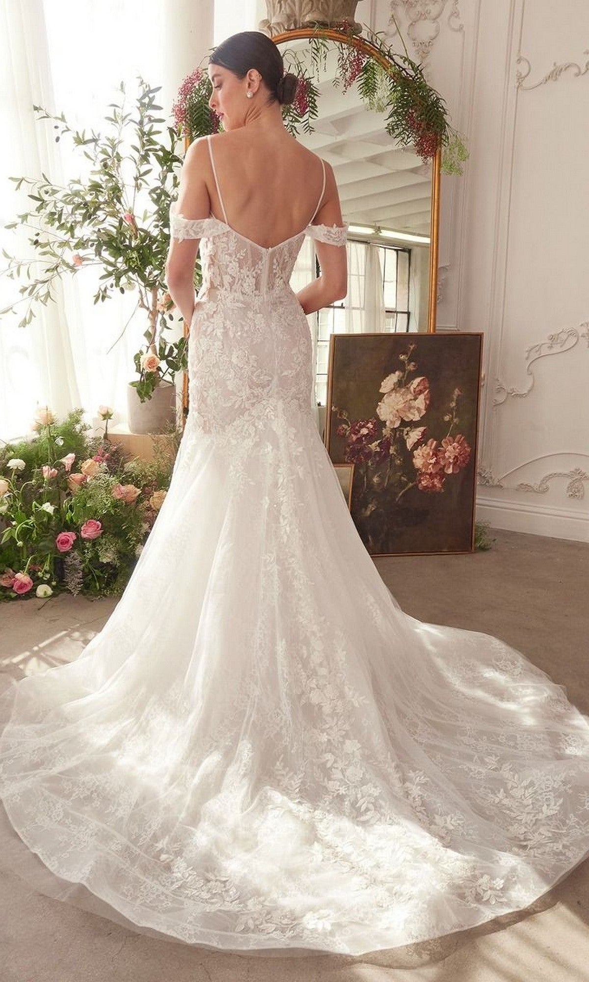 A woman in the Andrea and Leo WL051 long wedding dress with lace applique stands in a bright room with floral decor and a flower painting on the floor.